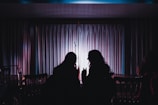 Two silhouetted people are seated in front of a stage with a microphone stand. The stage is framed by heavy curtains that are illuminated by soft pink and purple lighting, creating a moody and intimate ambiance. One person is holding a drink, suggesting a casual and comfortable setting.