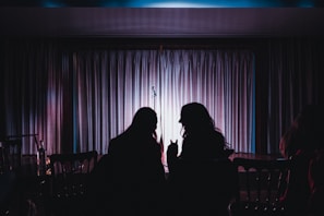 Two silhouetted people are seated in front of a stage with a microphone stand. The stage is framed by heavy curtains that are illuminated by soft pink and purple lighting, creating a moody and intimate ambiance. One person is holding a drink, suggesting a casual and comfortable setting.