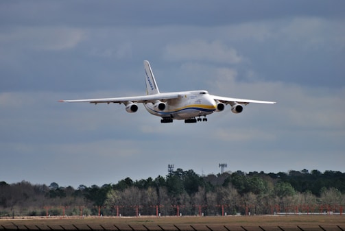 A large cargo aircraft flying low in the sky, approaching a runway for landing. The backdrop features a cloudy sky and a tree line at the edge of a clearing. The aircraft is prominent and centrally positioned, indicating its massive size.