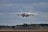 A large cargo aircraft flying low in the sky, approaching a runway for landing. The backdrop features a cloudy sky and a tree line at the edge of a clearing. The aircraft is prominent and centrally positioned, indicating its massive size.