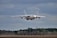 A large cargo aircraft flying low in the sky, approaching a runway for landing. The backdrop features a cloudy sky and a tree line at the edge of a clearing. The aircraft is prominent and centrally positioned, indicating its massive size.