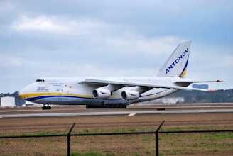 A large cargo airplane is taxiing on a runway at an airport. The aircraft is white with blue and yellow accents and has the word 'ANTONOV' displayed prominently on its tail fin. The background features a cloudy sky and some distant airport infrastructure.