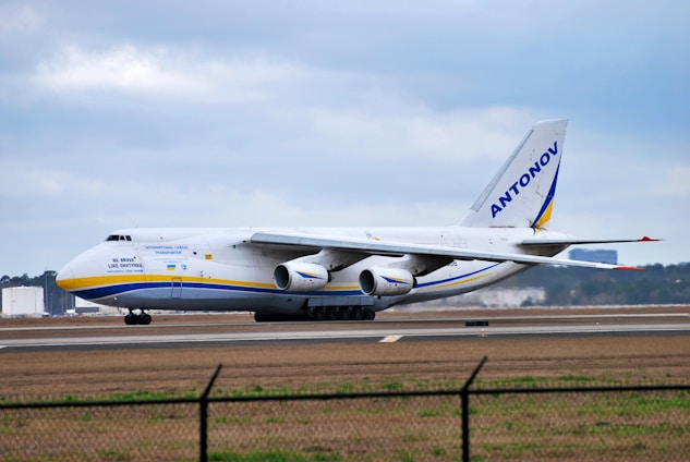 A large cargo airplane is taxiing on a runway at an airport. The aircraft is white with blue and yellow accents and has the word 'ANTONOV' displayed prominently on its tail fin. The background features a cloudy sky and some distant airport infrastructure.