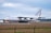 A large cargo airplane is taxiing on a runway at an airport. The aircraft is white with blue and yellow accents and has the word 'ANTONOV' displayed prominently on its tail fin. The background features a cloudy sky and some distant airport infrastructure.
