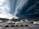 A parking lot in a commercial area is under a dramatic sky dominated by dark, swirling storm clouds. Several cars are parked, and trees surround the edges of the lot. The approaching storm contrasts with the stationary scene of parked vehicles, adding a sense of tension.