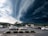 A parking lot in a commercial area is under a dramatic sky dominated by dark, swirling storm clouds. Several cars are parked, and trees surround the edges of the lot. The approaching storm contrasts with the stationary scene of parked vehicles, adding a sense of tension.