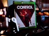 A video game case for the Xbox One sits prominently on a dark surface. The game is titled 'Control' and features a dramatic cover image of a character in a dynamic pose with a red and black color scheme. Surrounding the game case are various electronic gadgets and accessories, including headphones, coins, and a coiled cable.