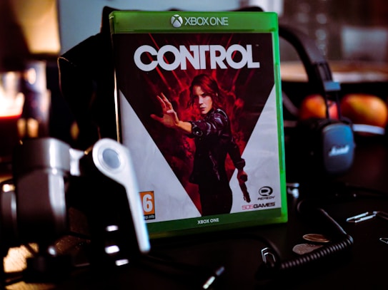 A video game case for the Xbox One sits prominently on a dark surface. The game is titled 'Control' and features a dramatic cover image of a character in a dynamic pose with a red and black color scheme. Surrounding the game case are various electronic gadgets and accessories, including headphones, coins, and a coiled cable.