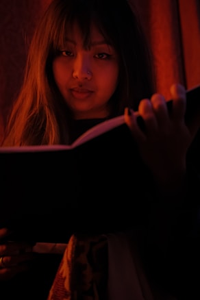 A person is holding an open book, gazing directly at the viewer. The lighting is warm and dim, creating an intimate and mysterious atmosphere. Long hair frames the face, and the image is dominated by deep shadows and rich, red tones.