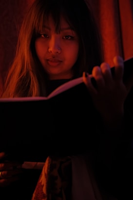 A person is holding an open book, gazing directly at the viewer. The lighting is warm and dim, creating an intimate and mysterious atmosphere. Long hair frames the face, and the image is dominated by deep shadows and rich, red tones.