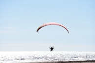 A person is engaged in powered paragliding over a body of water with a bright and clear sky above. The paraglider is silhouetted against the reflective surface of the sea, with the parachute displaying a blend of red and yellow.