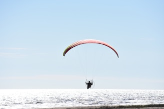 A person is engaged in powered paragliding over a body of water with a bright and clear sky above. The paraglider is silhouetted against the reflective surface of the sea, with the parachute displaying a blend of red and yellow.