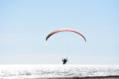 A person is engaged in powered paragliding over a body of water with a bright and clear sky above. The paraglider is silhouetted against the reflective surface of the sea, with the parachute displaying a blend of red and yellow.