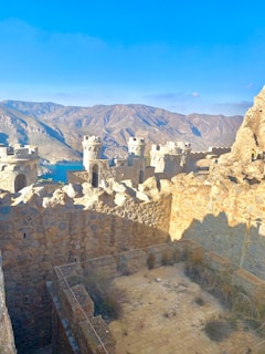 A stone fortress with turrets situated in a mountainous landscape. The fortifications are made of rugged, sandy-colored stones, and the surrounding mountains are vast and arid, with a clear blue sky above. Some grass and vegetation are growing in the courtyard area.
