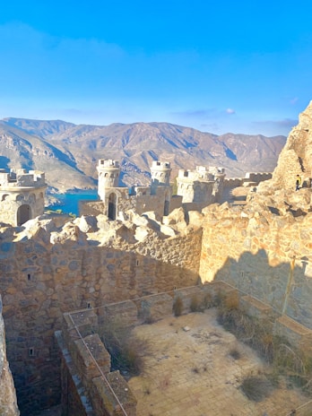 A stone fortress with turrets situated in a mountainous landscape. The fortifications are made of rugged, sandy-colored stones, and the surrounding mountains are vast and arid, with a clear blue sky above. Some grass and vegetation are growing in the courtyard area.