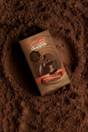 A box of cocoa powder is placed on a surface covered with loose cocoa powder, creating a rich, textured background. The packaging features graphic elements and text in another language.