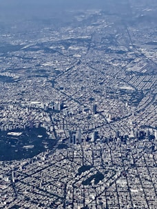 An aerial view of a sprawling urban cityscape with dense building coverage and complex street grid patterns. Skyscrapers and high-rise buildings are concentrated in certain areas, along with patches of greenery interspersed throughout the city.