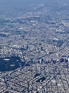 An aerial view of a sprawling urban cityscape with dense building coverage and complex street grid patterns. Skyscrapers and high-rise buildings are concentrated in certain areas, along with patches of greenery interspersed throughout the city.