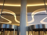 A modern architectural interior featuring sleek, curved ceiling lights and large glass panels. The lighting is warm, creating elegant reflections on the metallic surfaces of the revolving doors below.