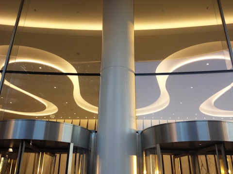 A modern architectural interior featuring sleek, curved ceiling lights and large glass panels. The lighting is warm, creating elegant reflections on the metallic surfaces of the revolving doors below.