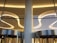 A modern architectural interior featuring sleek, curved ceiling lights and large glass panels. The lighting is warm, creating elegant reflections on the metallic surfaces of the revolving doors below.