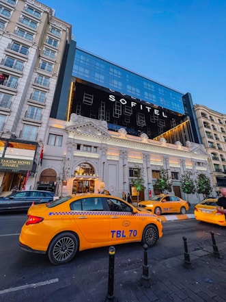 A modern hotel building with large reflective windows and ornate architectural details at the entrance. Several bright yellow taxis are parked on the street in front of the hotel. The scene is set in an urban environment with a clear blue sky overhead.