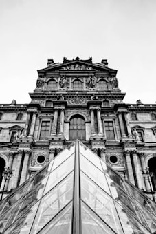 A black and white photograph featuring a classic architectural fa&ccedil;ade with intricate sculptures and columns. In the foreground, the geometric lines of a glass pyramid are prominently displayed, highlighting a blend of modern and historical architectural styles.