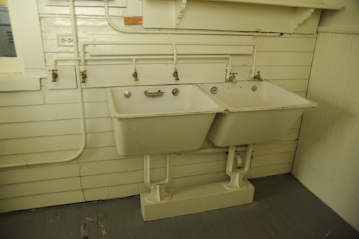 A pair of vintage double basins or utility sinks is mounted on a wall with several exposed pipes and faucets above them. The sinks and surrounding area are aged, with some discoloration on the basins and slight wear and tear on the wooden wall paneling. The room has a muted, utilitarian appearance with a grayish tone on the floor and off-white coloring on the walls and sinks.