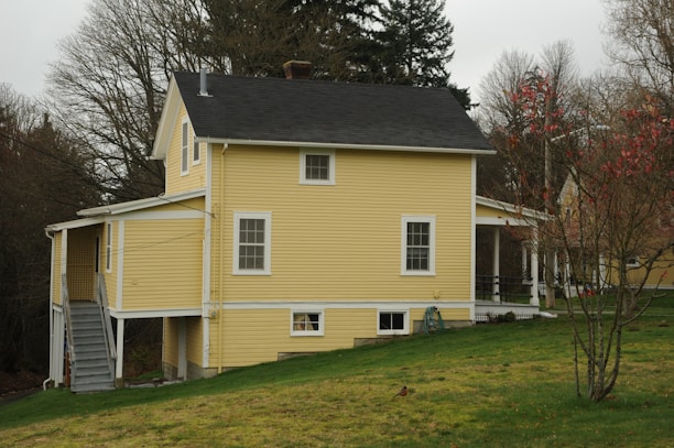 A two-story yellow house with a black roof situated on a grassy hill. The house has a side staircase leading up to an entrance, and a porch with white railings. Bare trees surround the property, suggesting autumn or winter season.