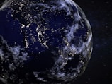 A view of Earth from space during the night, with illuminated city lights visible across various regions, highlighting population centers. The planet is partially covered by clouds, and the stars and distant galaxies are visible in the background.