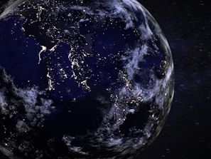 A view of Earth from space during the night, with illuminated city lights visible across various regions, highlighting population centers. The planet is partially covered by clouds, and the stars and distant galaxies are visible in the background.