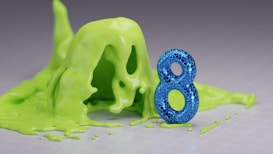 A blue, glittery number eight stands upright on a surface, surrounded by a puddle of bright green slime or liquid that appears to be spreading around it.