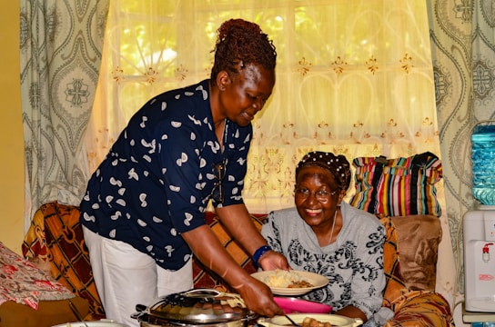 An elderly woman with a joyful expression receives a plate of food from a younger woman who is smiling warmly. They are indoors, surrounded by patterned curtains, a sofa with colorful cushions, and a water dispenser in the background.