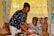 An elderly woman with a joyful expression receives a plate of food from a younger woman who is smiling warmly. They are indoors, surrounded by patterned curtains, a sofa with colorful cushions, and a water dispenser in the background.