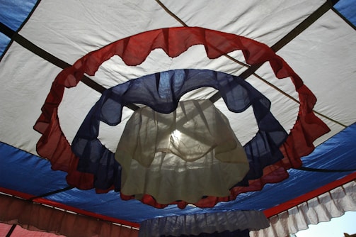 A fabric drapery with concentric circles of translucent material in shades of red, blue, and beige creates a layered effect. The colors are arranged in a circular pattern above, with light filtering through, casting subtle shadows.