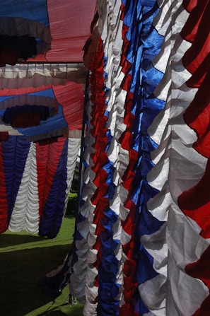 Colorful fabric drapes in red, white, and blue are hanging in a layered pattern. The fabric appears to be part of a decoration or tent setup, with pleated and ruffled textures creating a festive atmosphere. Sunlight accentuates the vibrant colors and shadows of the fabric.