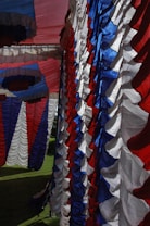 Colorful fabric drapes in red, white, and blue are hanging in a layered pattern. The fabric appears to be part of a decoration or tent setup, with pleated and ruffled textures creating a festive atmosphere. Sunlight accentuates the vibrant colors and shadows of the fabric.