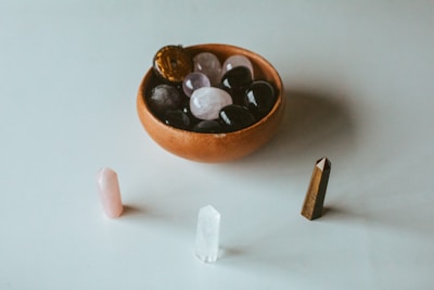 A collection of polished stones and crystals is arranged, featuring a small bowl filled with smooth, rounded dark and light stones, accompanied by three upright crystal points in pink, clear, and brown colors on a white surface.