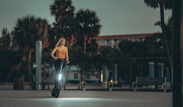 A person rides an electric scooter along a paved pathway surrounded by benches and tall palm trees. The background includes a building with balconies, set against a dusky, dimly lit sky. The lighting is soft and the scene appears to be taken during early morning or late evening.