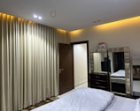 A neatly arranged bedroom featuring a large bed with white linens in the foreground. Alongside the bed is a mirror with a padded frame, situated on top of a dark-colored dresser. The walls are adorned with floor-to-ceiling beige curtains. There is a wooden door slightly ajar, leading to a brightly lit hallway.