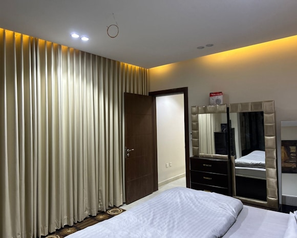 A neatly arranged bedroom featuring a large bed with white linens in the foreground. Alongside the bed is a mirror with a padded frame, situated on top of a dark-colored dresser. The walls are adorned with floor-to-ceiling beige curtains. There is a wooden door slightly ajar, leading to a brightly lit hallway.