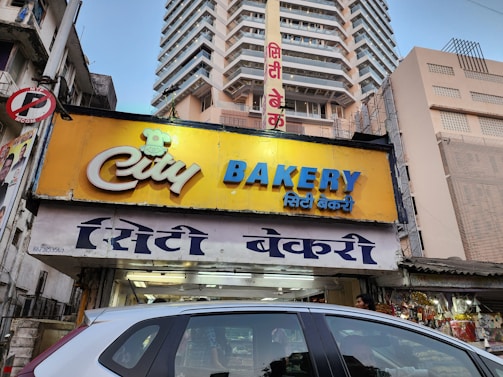 A bakery storefront sign with a yellow background and blue and white text. The bakery is located on a busy street with several tall buildings in the background. The shop's name is prominently displayed in both English and Hindi. A car is partially visible in the foreground, and there are various shop items visible through the storefront.