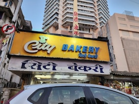 A bakery storefront sign with a yellow background and blue and white text. The bakery is located on a busy street with several tall buildings in the background. The shop's name is prominently displayed in both English and Hindi. A car is partially visible in the foreground, and there are various shop items visible through the storefront.