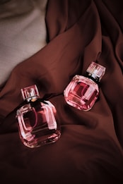 Two pink crystal perfume bottles with black ribbon details are placed on a rich brown fabric. The fabric drapes elegantly, providing a luxurious backdrop that complements the elegance of the bottles. The lighting highlights the glossy and reflective surfaces of the bottles.