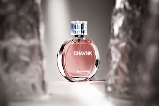 A perfume bottle with a pink liquid stands elegantly in soft lighting. The bottle has a transparent cap and sleek design with reflective surfaces, positioned between blurred translucent objects.