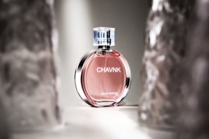 A perfume bottle with a pink liquid stands elegantly in soft lighting. The bottle has a transparent cap and sleek design with reflective surfaces, positioned between blurred translucent objects.
