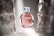 A perfume bottle with a pink liquid stands elegantly in soft lighting. The bottle has a transparent cap and sleek design with reflective surfaces, positioned between blurred translucent objects.