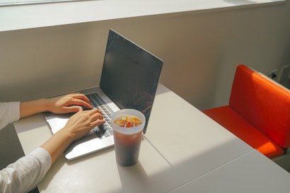 A person is typing on a laptop placed on a white table next to a window. A large plastic cup filled with iced coffee is positioned to the right of the laptop. Sunlight streams in from the window, casting shadows on the table. An orange chair is visible on the right side of the image.