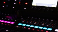 A close-up view of a sound mixing console featuring rows of illuminated buttons and dials. The console has multiple colored buttons, including pink, blue, and red, arranged in sequences. A screen is partially visible in the upper right corner of the frame, displaying technical information. The ambient lighting is dim, highlighting the bright colors of the control panel.
