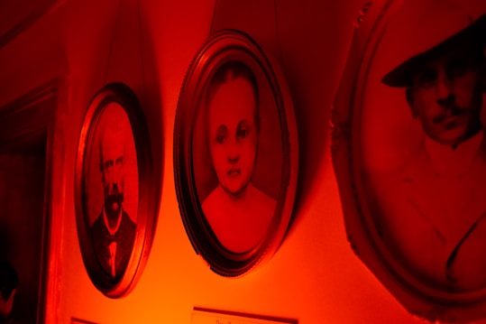 Three vintage portraits are hanging on a wall. The image is bathed in a deep red light, creating an almost eerie atmosphere. The center portrait features a young child with a neutral expression, and the portraits on either side depict adults with stern looks, possibly from the early 20th century, as suggested by their attire.